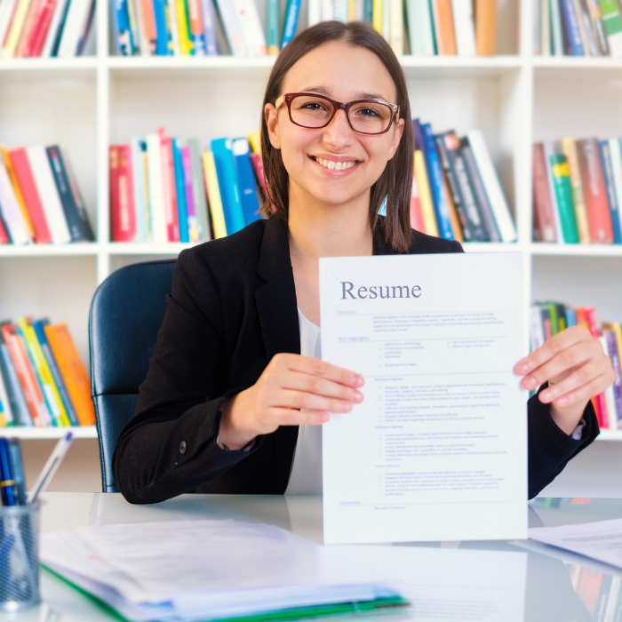 Women sitting in front of a bookshelf holding up a paper that says Resume at the top